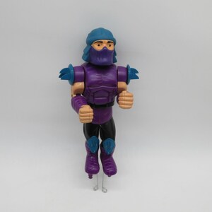May include: A vintage action figure of a character in a purple and black outfit with a blue helmet and mask. The figure has tan hands and forearms, and blue shoulder and knee pads. The figure is standing on a clear stand.