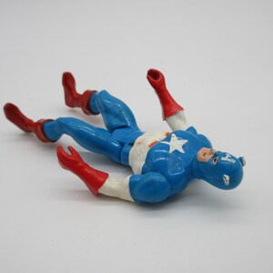 May include: A blue and red plastic action figure of Captain America lying on its back. The figure is wearing a blue and red costume with a white star on the chest. The figure has a red and white mask.