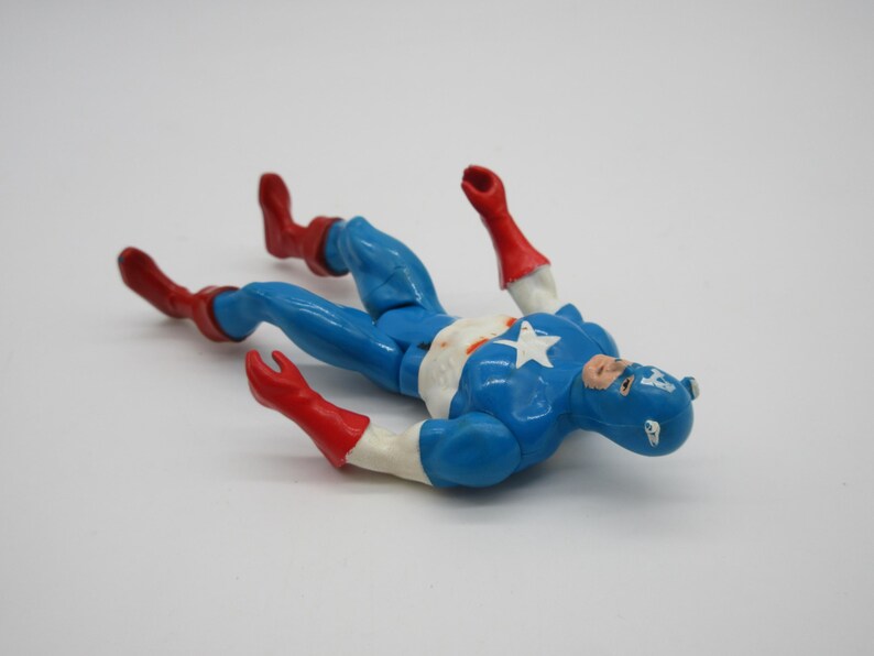 May include: A blue and red plastic action figure of Captain America lying on its back. The figure is wearing a blue and red costume with a white star on the chest. The figure has a red and white mask.