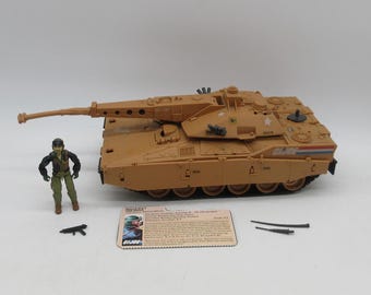 May include: Tan toy tank with a long barrel, a small action figure, and a card with text and a graphic. The tank has multiple details, including wheels and a turret. The action figure is wearing a green uniform. The card has the text "G.I. Joe".