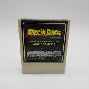May include: A brown and beige Atari 2600 video game cartridge with the title "Roc'n Rope" in yellow and red letters. The cartridge has a black label with white text that reads "Video Game Cartridge for use with Atari 2600 VCS" and copyright information for Konami Industry Co., Ltd. 1983.