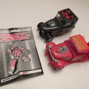 Könnte beinhalten: Zwei Transformers Generation 1 Spielzeuge in ihren Fahrzeugformen. Das rote Spielzeug ist ein Hot Rod mit goldenen Flammen auf dem Dach. Das schwarze Spielzeug ist ein Hot Rod mit einem schwarz-roten Schachbrettmuster auf dem Dach. Die Spielzeuge befinden sich auf einer weißen Oberfläche. Die Anleitung für das rote Spielzeug ist auf dem Bild zu sehen. Die Anleitung enthält den Text "Heroic Autobot" und "Volt".