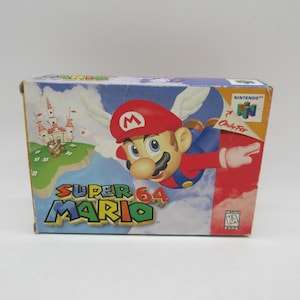 Puede incluir: Una caja de cartón rectangular de videojuego para Super Mario 64 para la consola Nintendo 64. El frente muestra a Mario volando con alas blancas, un castillo al fondo, el título y un símbolo de clasificación K-A sobre fondo blanco.