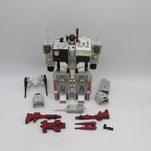 May include: A grey and white robot action figure with black and red accents, featuring a Decepticon emblem on the chest. The figure is accompanied by several detachable plastic accessories, including red blasters, small vehicle components, and grey armour pieces.