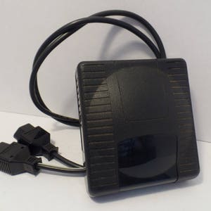 May include: A black foot pedal with a textured surface and a black cord with two plugs. The pedal has a black button on the top.