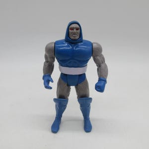 May include: A plastic action figure of a muscular character with grey skin, a blue chest piece, blue gloves, blue boots, and a white belt. The figure has a hooded head design with prominent red eyes and a stern facial expression, standing in a neutral pose.