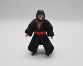 May include: A black action figure wearing a black robe with a red sash. The figure has a black beard and mustache and is wearing a black head covering.