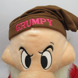 Puede incluir: Un peluche de Gruñón de Blancanieves. El juguete presenta un sombrero marrón con la palabra "GRUMPY" bordada en rosa. Gruñón tiene una expresión de mal humor con grandes ojos negros y una barba blanca.