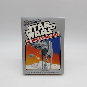Puede incluir: Una caja de cartucho de videojuego vintage de Star Wars: El Imperio Contraataca. La caja presenta el logotipo de Star Wars y una ilustración de un caminante AT-AT. El texto en la caja dice "A VIDEO GAME CARTRIDGE FROM PARKER BROTHERS" y "FOR ATARI & SEARS VIDEO GAME SYSTEMS."