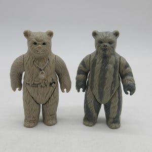 Op de afbeelding: Twee vintage Star Wars Ewok actiefiguren. Eén figuur is bruin met een gedetailleerd borststuk, de andere is grijs met strepen. Beide figuren staan rechtop en hebben een beerachtig uiterlijk.