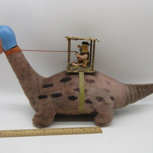 Puede incluir: Un juguete de peluche vintage de un dinosaurio marrón y negro con manchas y un casco azul. Una pequeña figura de plástico marrón de un hombre en una jaula se sienta en la espalda del dinosaurio.