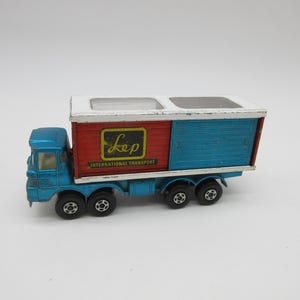 May include: A blue toy truck with a white cab and a red and blue box trailer. The trailer has the words "Lep International Transport" printed on the side.