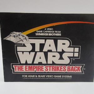 May include: A black and white box with a red and white logo for the Star Wars: The Empire Strikes Back video game cartridge for Atari and Sears video game systems. The box features a white spaceship illustration and the text "A Video Game Cartridge From Parker Brothers".