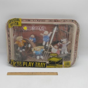 May include: A rectangular Cabbage Patch Kids TV and play tray featuring a farm scene with five dolls, a wooden wheel, and a pitchfork. The tray is wrapped in clear plastic with a yellow Special Offer sticker and a wooden ruler measuring 12 inches.