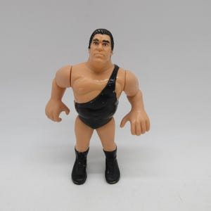 Könnte beinhalten: Eine Kunststoff-Actionfigur eines Ringers mit gebräuntem Teint, schwarzen Haaren und muskulösem Körperbau. Die Figur trägt ein schwarzes Wrestling-Singlet mit einer Schulter und schwarze Schnürstiefel. Die Arme sind seitlich ausgestreckt.