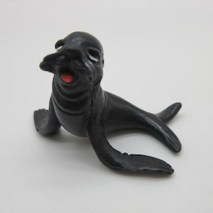 May include: A black plastic toy seal with a red mouth and white eyes. The seal is sitting on a white surface.