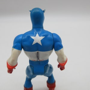 May include: A blue and white plastic action figure of Captain America. The figure is standing with its back to the camera. The figure has a red star on its chest and a red stripe across its waist.
