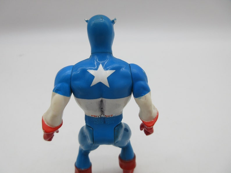 May include: A blue and white plastic action figure of Captain America. The figure is standing with its back to the camera. The figure has a red star on its chest and a red stripe across its waist.