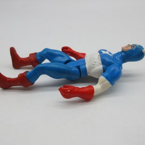 May include: A vintage action figure of Captain America lying on its back. The figure is made of plastic and is painted in the classic red, white, and blue colors. The figure has a red and blue costume with a white star on the chest. The figure has a red mask and a white star on the forehead.