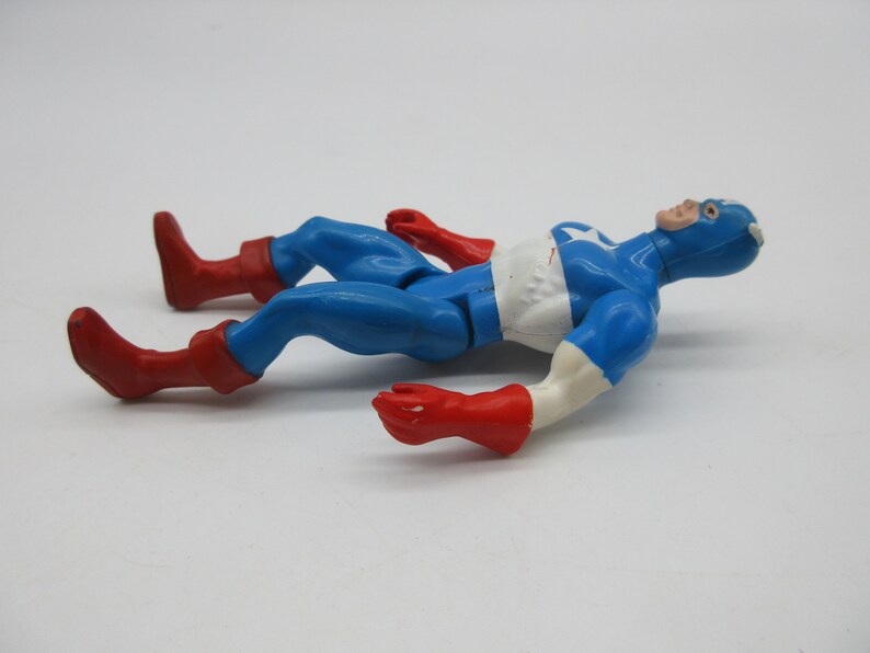 May include: A vintage action figure of Captain America lying on its back. The figure is made of plastic and is painted in the classic red, white, and blue colors. The figure has a red and blue costume with a white star on the chest. The figure has a red mask and a white star on the forehead.