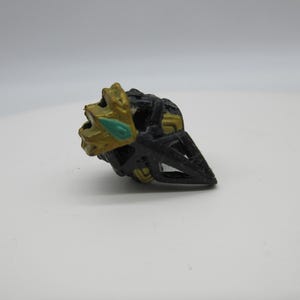 Op de afbeelding: Een zwart en gouden plastic speelgoed met een groene edelsteen. Het speelgoed is gevormd als een helm of hoofd.