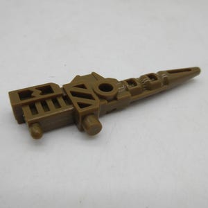 May include: A brown plastic toy accessory featuring a long, tapered barrel with a pointed tip, a central circular opening, and a rectangular base with vertical slats and geometric cutouts, designed as a component for a small action figure.
