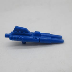 May include: A small, blue plastic toy weapon accessory featuring a double-barreled design with a raised central fin and a protruding peg on the underside. The object has a molded, futuristic aesthetic with geometric detailing along the main body.