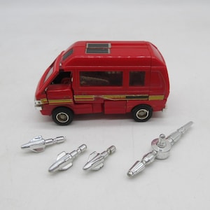 May include: A red toy van with black wheels and yellow and black side stripes, featuring a roof hatch. Beside the van are four separate silver-coloured plastic accessories, including three small pointed projectiles and one longer rod-like piece.