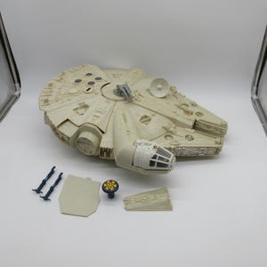 Op de afbeelding: Een wit plastic model van het ruimteschip Millennium Falcon uit Star Wars. Het model is gedemonteerd en bevat verschillende onderdelen, waaronder de romp, een afneembare cockpit en twee landingsgestellen.
