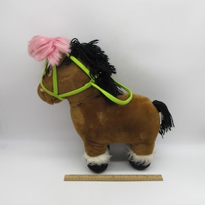 May include: Brown plush horse with a pink mane and tail, black mane and tail, and a green bridle. The horse is standing on a white surface.