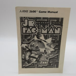 Puede incluir: Manual de juego en blanco y negro para el juego Atari 2600 Jr. Pac-Man. La portada presenta una imagen de dibujos animados de Jr. Pac-Man montando un triciclo.