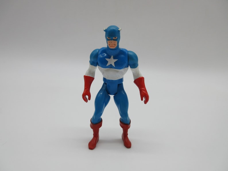 May include: A blue and red plastic action figure of Captain America. The figure has a white star on its chest and is wearing a blue helmet with a red stripe.