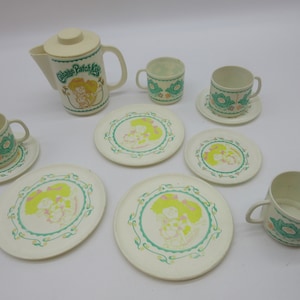 Op de afbeelding: Een vintage Cabbage Patch Kids theeservies van crèmekleurig plastic. De set bevat een theepot, kopjes, schotels en borden. Elk stuk heeft een Cabbage Patch Kids-afbeelding en bloemaccenten in groen, roze en geel.