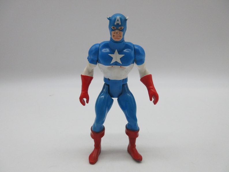 May include: A vintage action figure of Captain America, wearing his iconic red, white, and blue costume. The figure has a red helmet with a white star and the letter "A" on the front. The figure is standing with its arms at its sides.