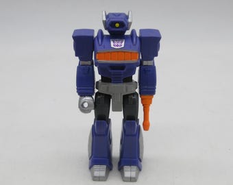 May include: A blue and orange robot action figure with a purple emblem. The robot has a silver arm and feet, and an orange arm with a gun. The figure is standing upright. The robot is approximately 15 cm tall.