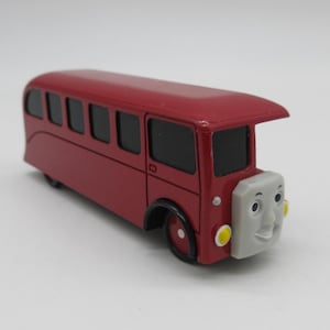 May include: A red toy bus with a grey front face featuring a smiling expression. The bus has black windows, yellow circular headlights, and black wheels. The design includes a prominent roof overhang above the front windscreen and a smooth finish.