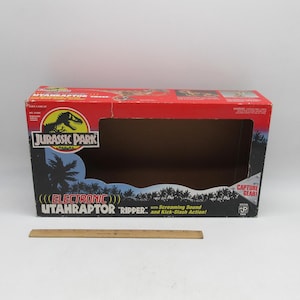 Op de afbeelding: Een rechthoekige rood-zwarte kartonnen speelgoedverpakking voor een Jurassic Park Electronic Utahraptor Ripper. De doos heeft een groot uitgesneden venster, palmbomengraphics en tekst waaronder "Screaming Sound and Kick-Slash Action". Een houten liniaal van 30 cm ligt ervoor.