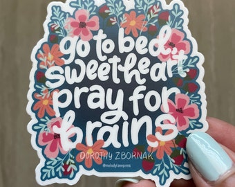 May include: A sticker with a floral border and the text "go to bed sweetheart pray for brains" in white lettering. The sticker is on a blue background and has the artist's name and social media handle at the bottom.