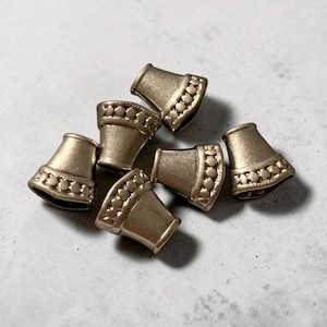 May include: A collection of small, metallic gold-colored bell-shaped beads. Each bead features a decorative dotted pattern around the top edge. The beads are scattered on a light gray surface.