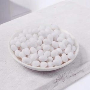 May include: A small, white ceramic dish filled with numerous white, heart-shaped candies. The dish sits on a textured, white surface, with a soft, neutral background. The candies appear to be sugared or coated.