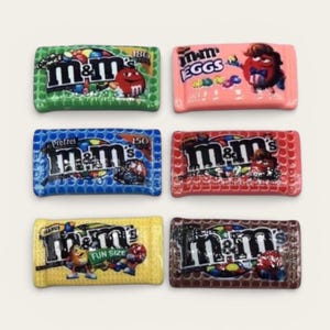 May include: Six rectangular M&M's candy package charms in green, pink, blue, red, yellow, and brown. Each charm features the M&M's logo, colorful candy illustrations, and specific variety text including Eggs, Pretzel, and Fun Size printed on the front.