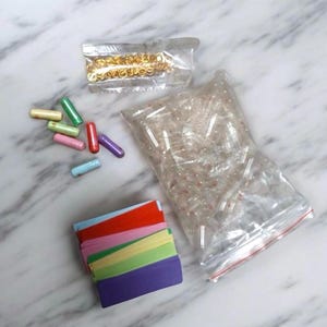 May include: A craft kit containing a large clear plastic bag filled with empty clear capsules, a smaller bag of gold-tone metal jump rings, several loose pastel-colored capsule charms, and a stack of rectangular paper strips in various colors.