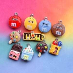 May include: A collection of colorful M&M's themed charms. The charms include various M&M's characters, ice cream bars, and a bag of M&M's. The charms are red, yellow, blue, and brown with the letter "m" on them.
