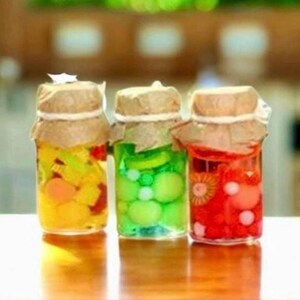 May include: Three clear glass jars filled with colorful round candies in yellow, green, and red. Each jar is topped with a brown paper lid cover secured by a thin white string tied around the neck, creating a uniform presentation of the sweets.