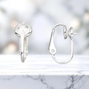 May include: Two silver-colored clip-on earrings. One earring is closed, showing the clip mechanism, while the other is open, displaying the hook and post design. The earrings are set against a blurred, light-colored background, resting on a white marble surface.
