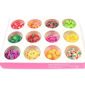 May include: A set of twelve small, clear jars filled with colorful fruit-shaped polymer clay slices. The fruit slices include strawberries, watermelon, kiwi, lemons, bananas, and grapes. The jars are arranged in a white box with a pink border.