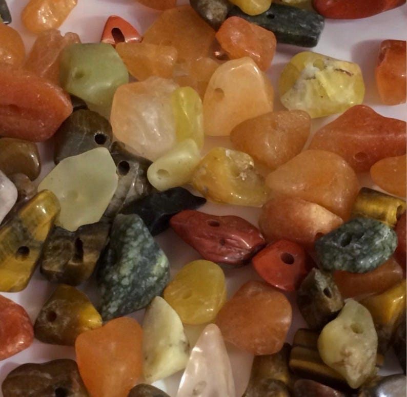 May include: A collection of polished gemstones in various colors, including orange, yellow, green, and red. The stones have irregular shapes and are drilled with holes for stringing. The gemstones are suitable for jewelry making or crafting.