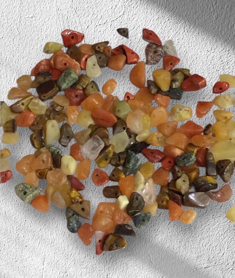 May include: A collection of small, colorful gemstone beads in shades of orange, yellow, red, and green. The beads are irregular in shape and have holes for stringing. The gemstones appear to be polished and have a natural, earthy look.