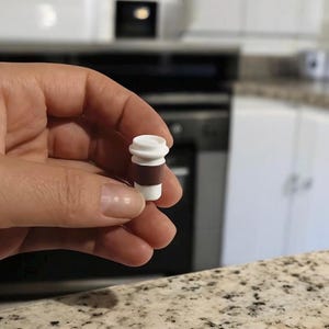 May include: A miniature coffee cup replica, featuring a white top and base with a brown midsection. The small cup is held in a hand, showcasing its detailed design. The background includes a kitchen setting with white cabinets and appliances.