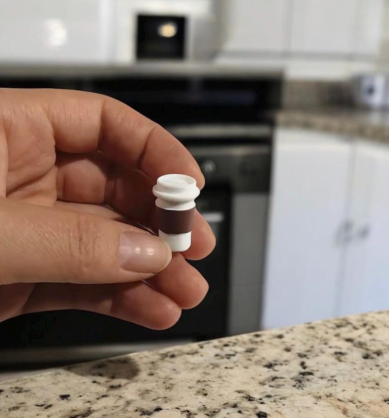 May include: A miniature coffee cup replica, featuring a white top and base with a brown midsection. The small cup is held in a hand, showcasing its detailed design. The background includes a kitchen setting with white cabinets and appliances.